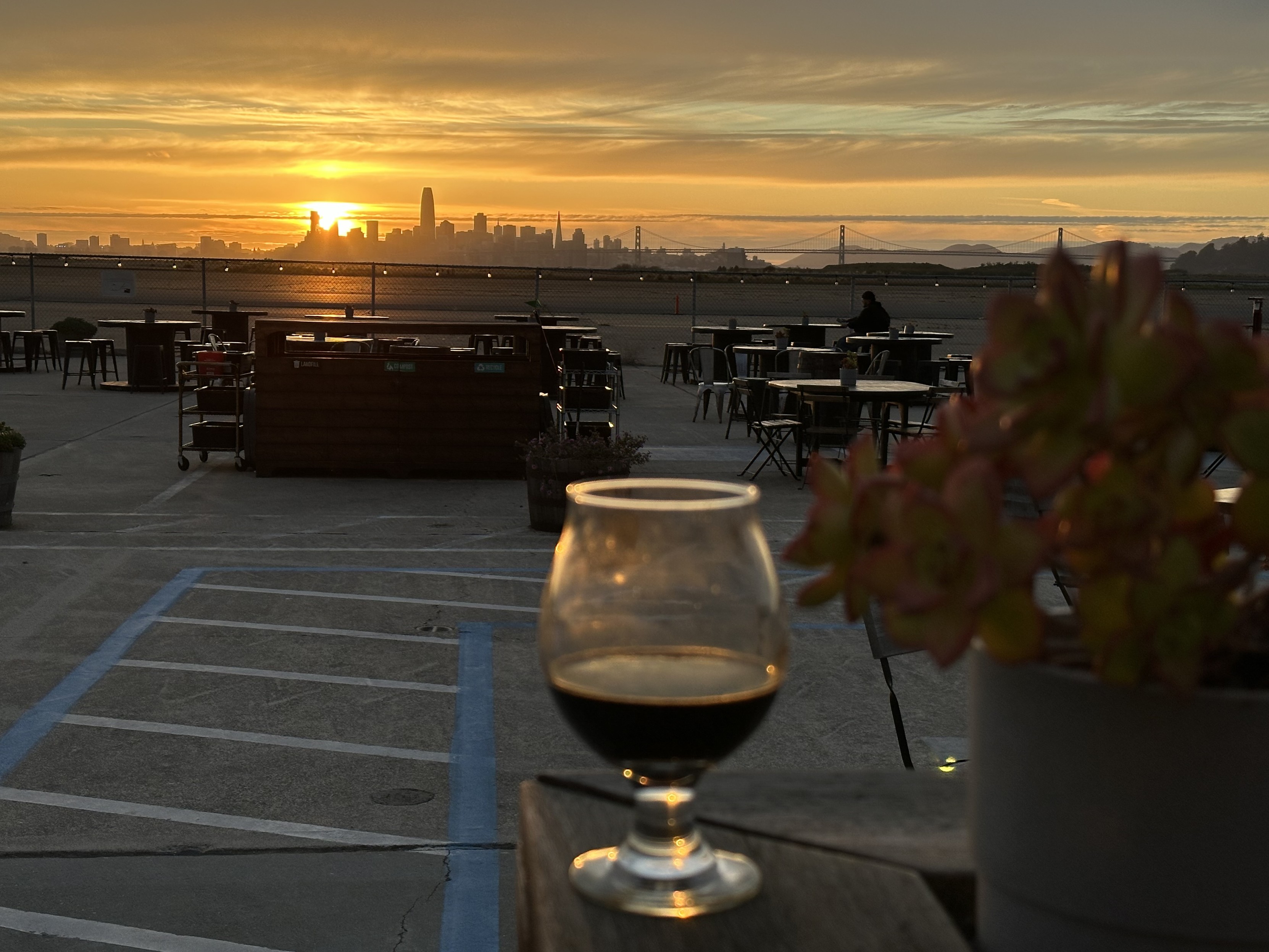 Small glass of beer out of focus in the front, in the back is the San Francisco skyline with the sun setting behind it.